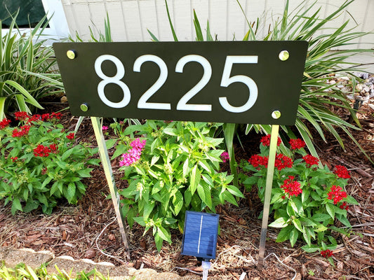 Solar Address numbers on a metal sign with yard stakes.  solar panel in front .  Sign and spotlight placed in ground in a garden.