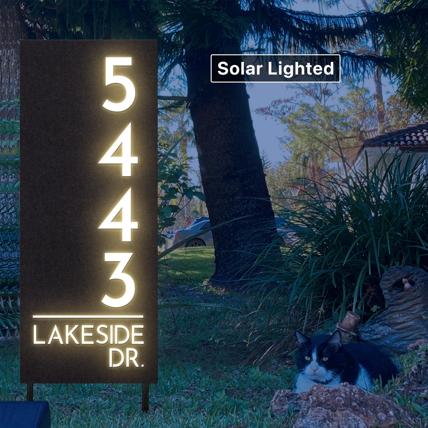 May include: A dark brown, solar-lighted house number sign displays the address 5443 Lakeside Dr.  The numbers are illuminated in bright white. The sign is mounted on a black metal stand and is situated in a garden setting with lush green grass and a black and white cat resting on a log in the foreground. The background includes trees and a portion of a house.  The words 'Solar Lighted' appear in a box above the sign.