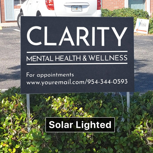 May include: A black sign with white text that reads "CLARITY MENTAL HEALTH & WELLNESS." Below, it says "For appointments" and provides a website and phone number. The sign is labeled "Solar Lighted" and is mounted on metal posts.